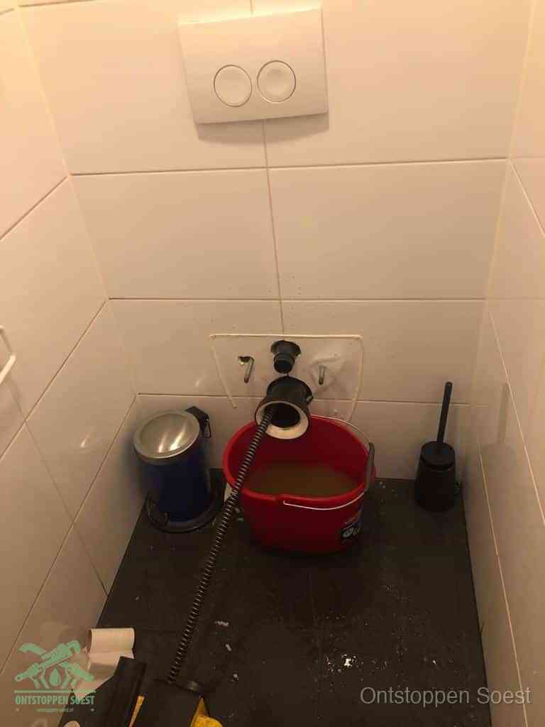 Een ontstopte wand-wc met verwijderde toiletpot waar een ontstoppingsveer in de wanduitgang verdwijnt, het achterhalen waar de verstopping zit in Soest