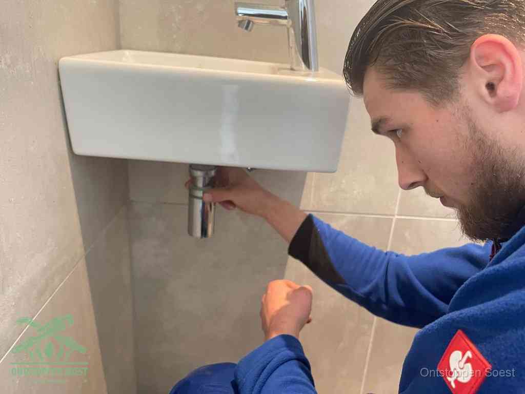 Een monteur draait met de hand de zwanenhalssifon onder een fonteinwasbak in de toiletruimte los om de bron van rioollucht in Soest te vinden