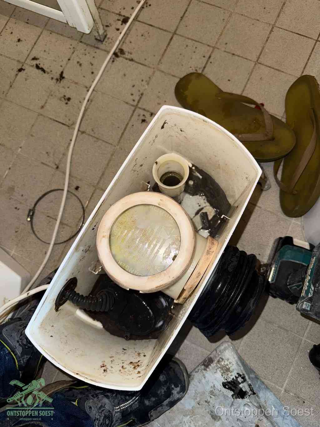 Een gedemonteerde toiletpot op de vloer met ontstoppingsveer en gereedschap ernaast, het verwijderen van een vastzittend voorwerp uit de wc in Soest
