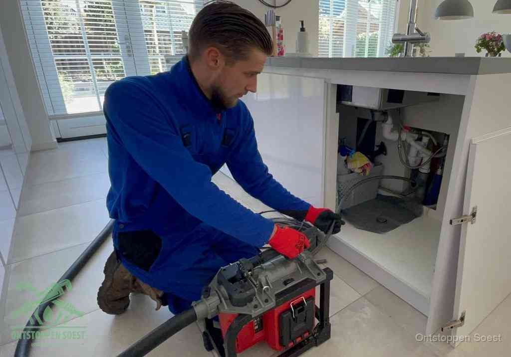 Een monteur voert een professionele elektrische ontstoppingsmachine in de afvoer onder de spoelbak tijdens het gootsteen ontstoppen in Soest